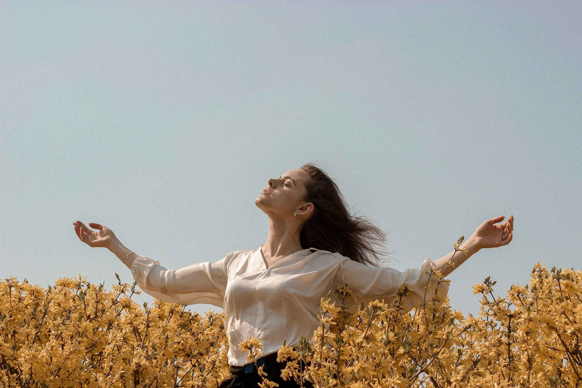 Woman with arms outstretched, eyes closed, in field of yellow flowers under blue sky.