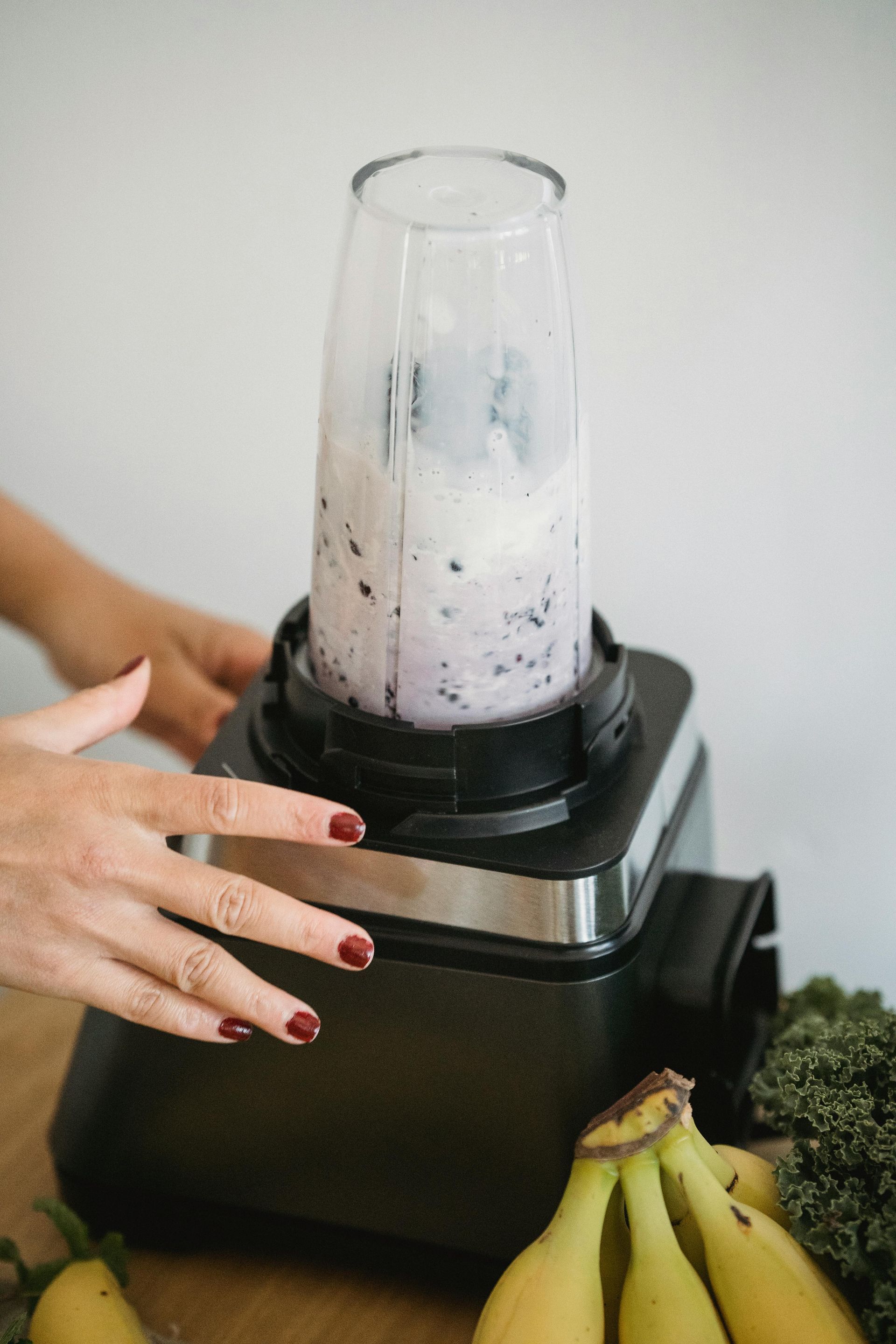 Blender with purple smoothie being prepared; bananas and greens in the foreground; woman's hands on blender.
