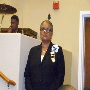 A person in a black blazer with a white lapel pin stands in front of a white podium, with a partially visible musician.
