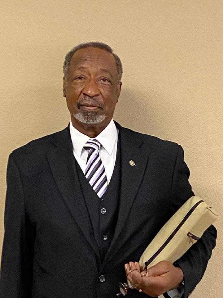 A man in a black suit, vest, and striped tie standing against a plain wall, holding a tan pouch in his hand.