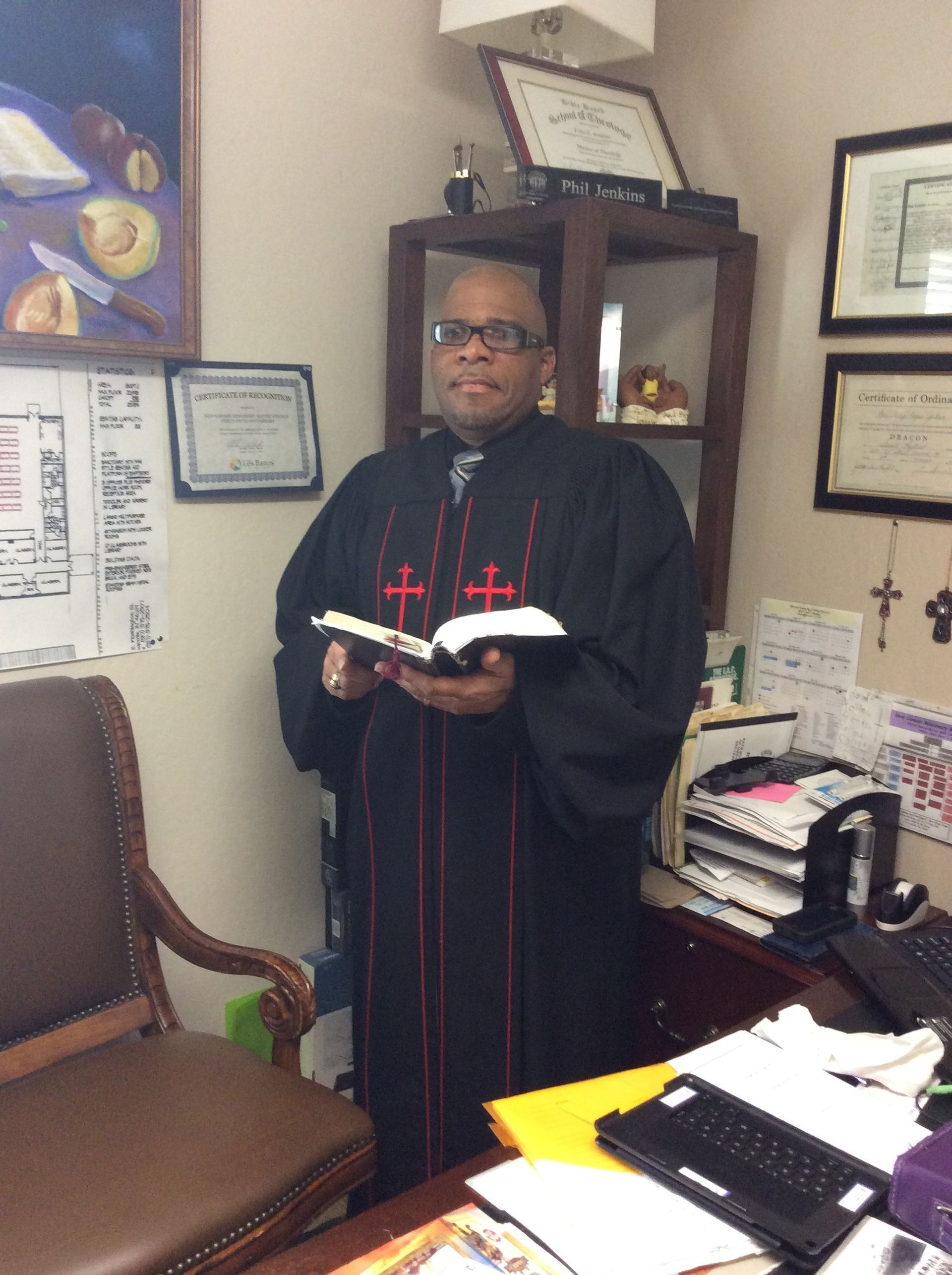 A person in clerical robes stands in an office holding an open book, with framed certificates on the wall behind them.