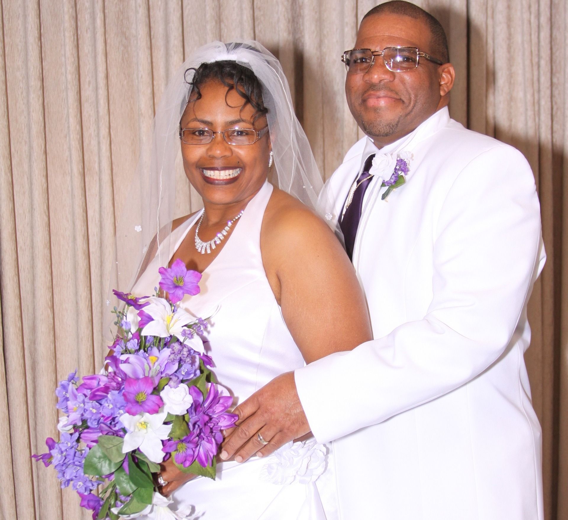 A bride in a white wedding dress and a groom in a white suit stand together, smiling, holding a purple and white bouquet.