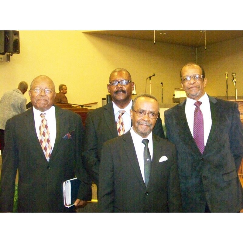 Four men in suits stand in a line against a neutral wall in what appears to be a formal indoor setting.