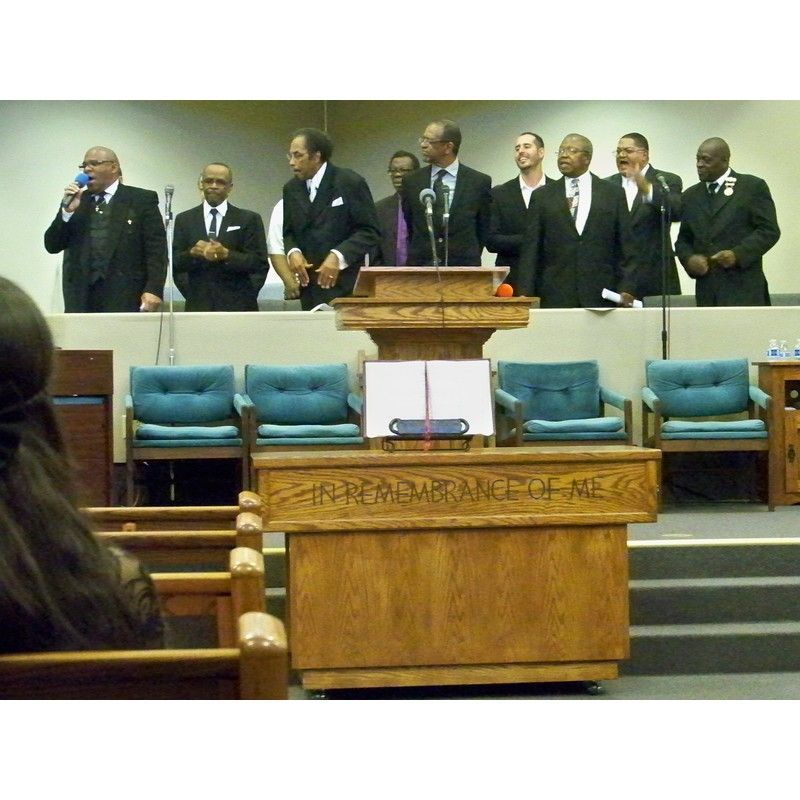 A group of men in suits stands behind a wooden pulpit inside a church, with one speaking into a microphone.
