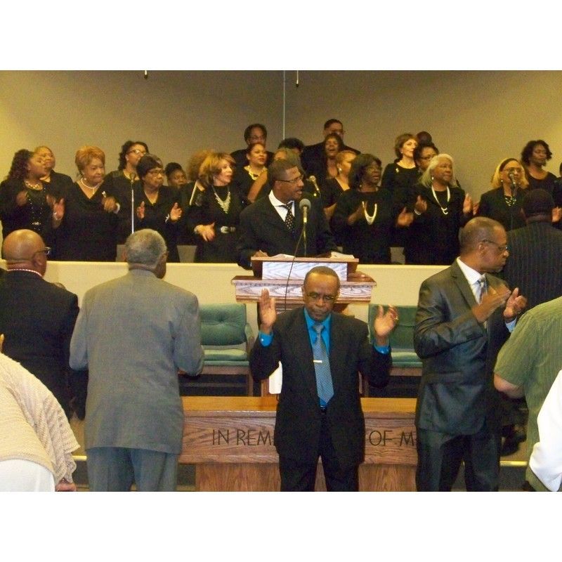 A choir stands behind a church altar while congregants stand and pray in the foreground during a service.