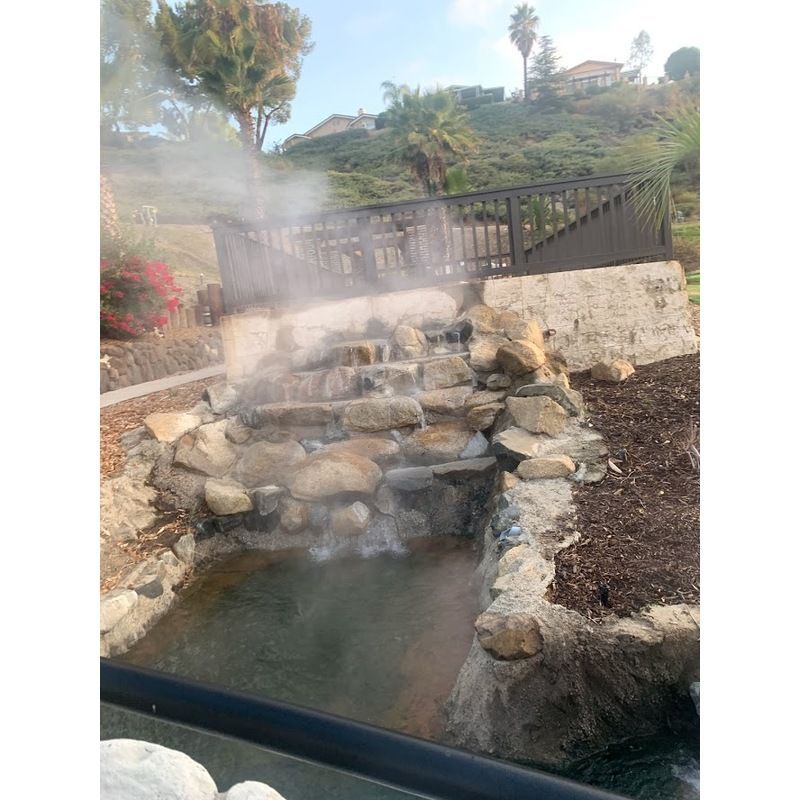 A steaming, terraced stone waterfall flowing into a small pond outdoors near a wooden deck and hillside.