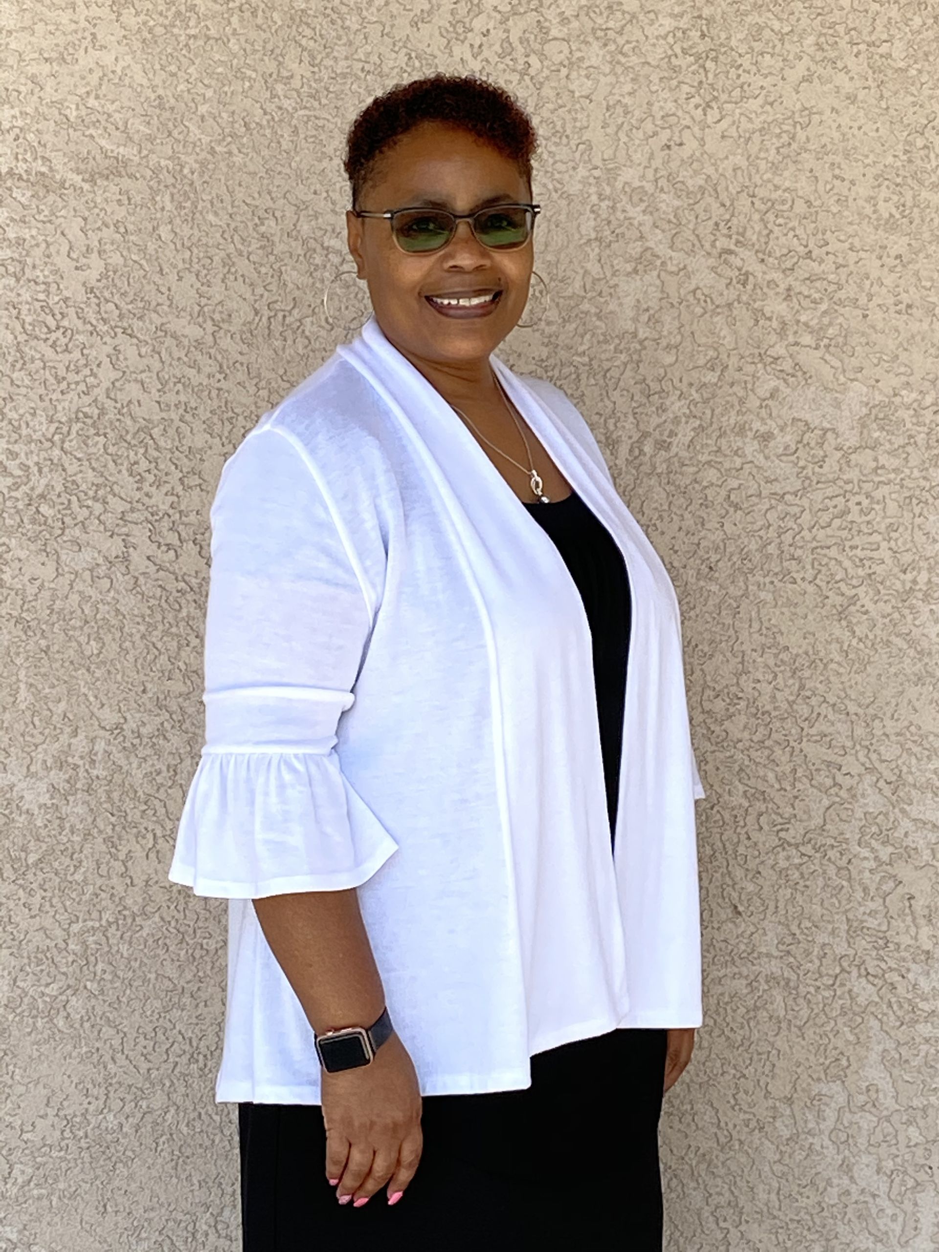 A person smiling, wearing a white, bell-sleeved cardigan and black top, stands before a textured, beige wall.
