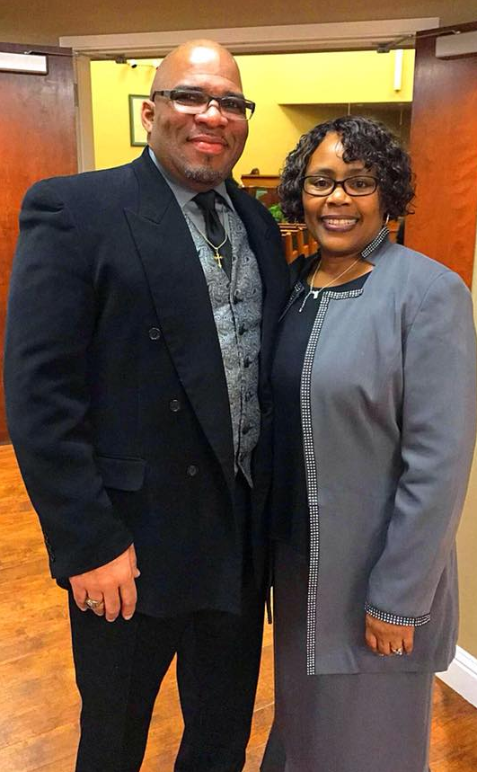 A man in a dark suit and a woman in a grey blazer standing side-by-side, smiling, in an indoor setting.