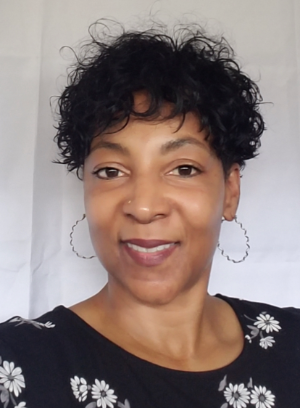 A person with short, dark, curly hair wearing hoop earrings and a black floral top, smiling against a white background.