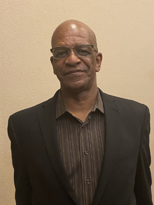 A person wearing glasses and a dark suit over a striped shirt stands against a plain beige wall.