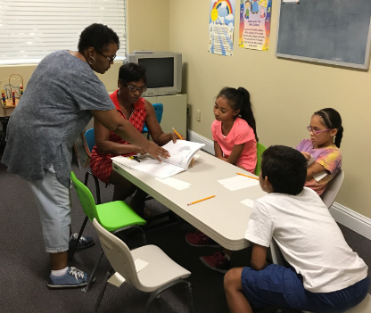 Two adults helping three children with schoolwork around a table in a classroom setting.