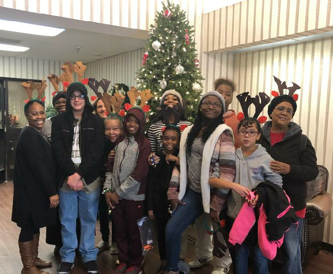 A group of people wearing reindeer antlers stand smiling in front of a decorated Christmas tree.