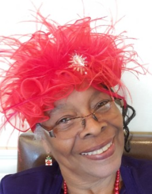 A smiling person wearing a red feathered hat, glasses, and a red beaded necklace against a plain background.