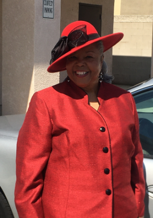 A smiling person wears a vibrant red suit and a matching red wide-brimmed hat with a dark decorative bow.