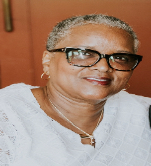 A person with short, silver hair and glasses smiles while wearing a white top and a thin gold necklace.