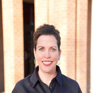 A person with short, dark hair and hoop earrings smiling, wearing a black collared shirt in front of a brick building.