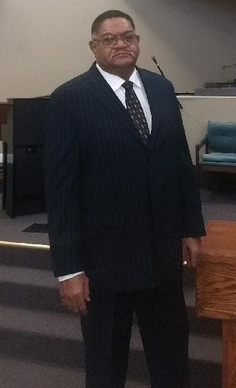 A person in a dark pinstripe suit and tie standing in what appears to be a church or formal assembly hall.