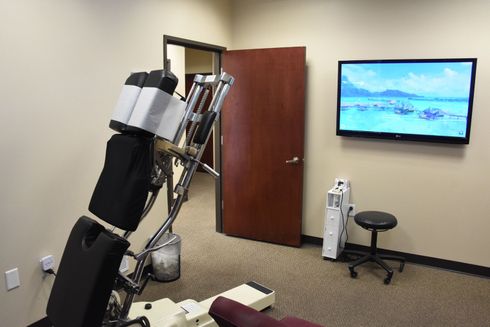 Chiropractic office interior: adjustable table, closed brown door, TV showing beach scene, small stool.