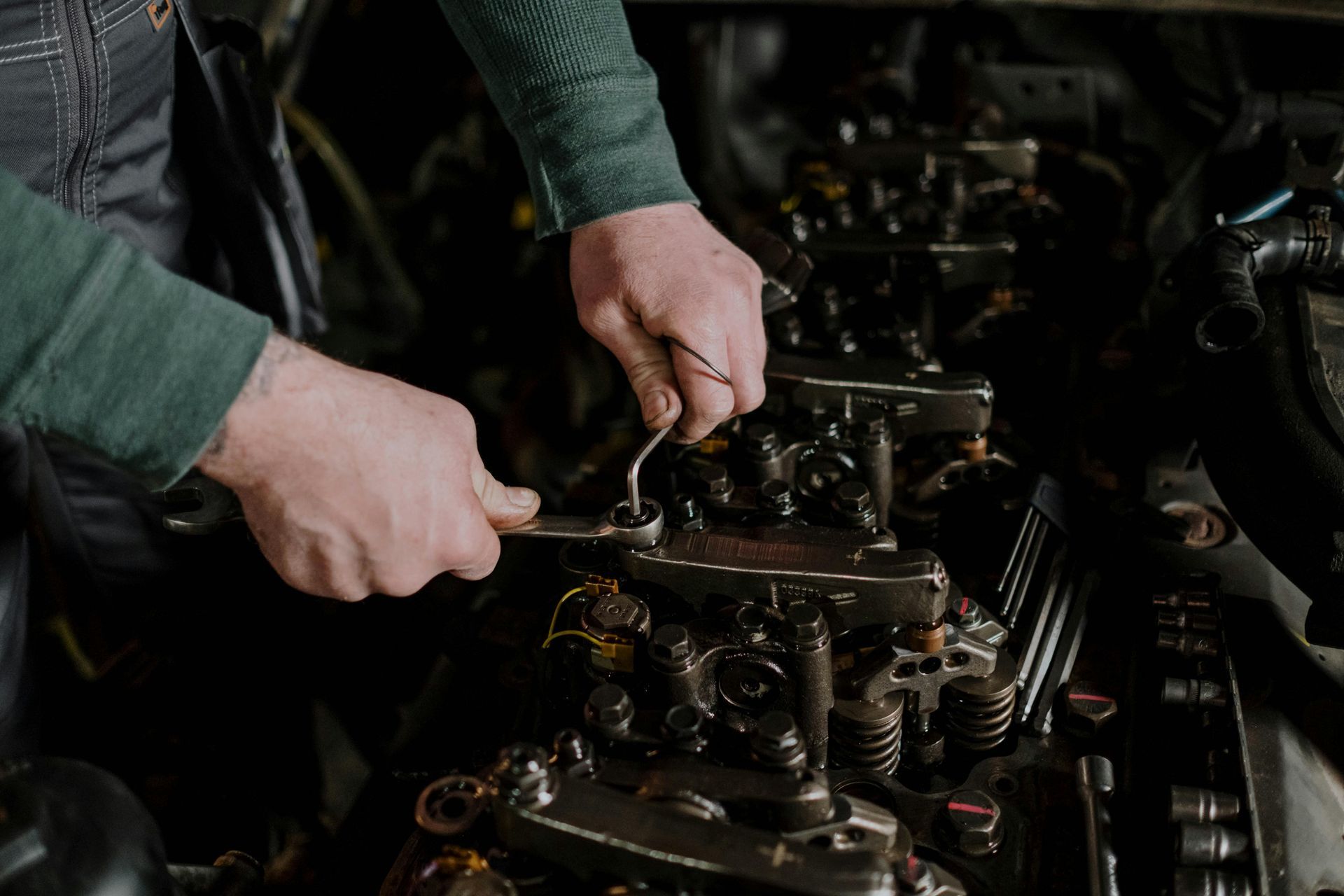 A person in a green shirt uses a wrench to adjust the mechanical components of an open car engine.