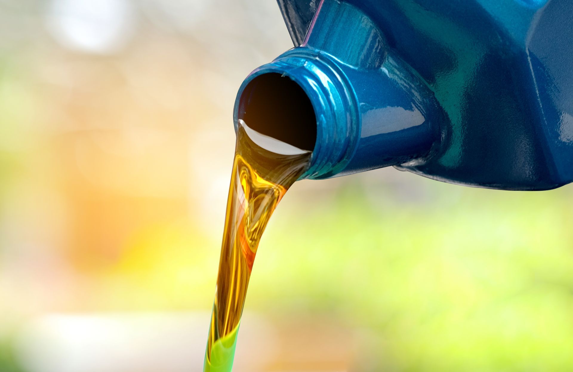 Golden motor oil pouring from a blue plastic container against a blurred, bright outdoor background.