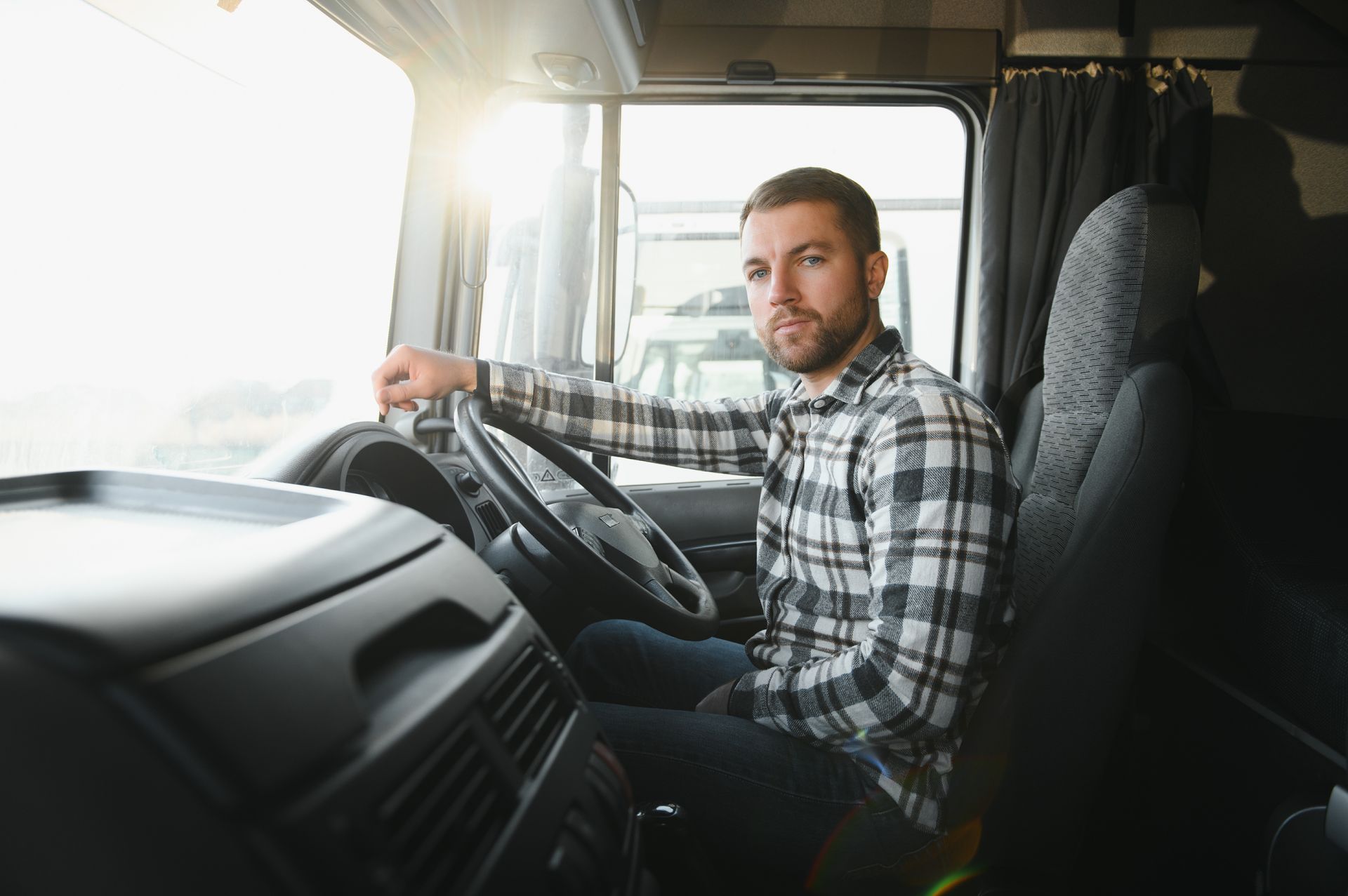 A person in a plaid shirt sits in the driver's seat of a truck, looking toward the camera with their hand on the wheel.