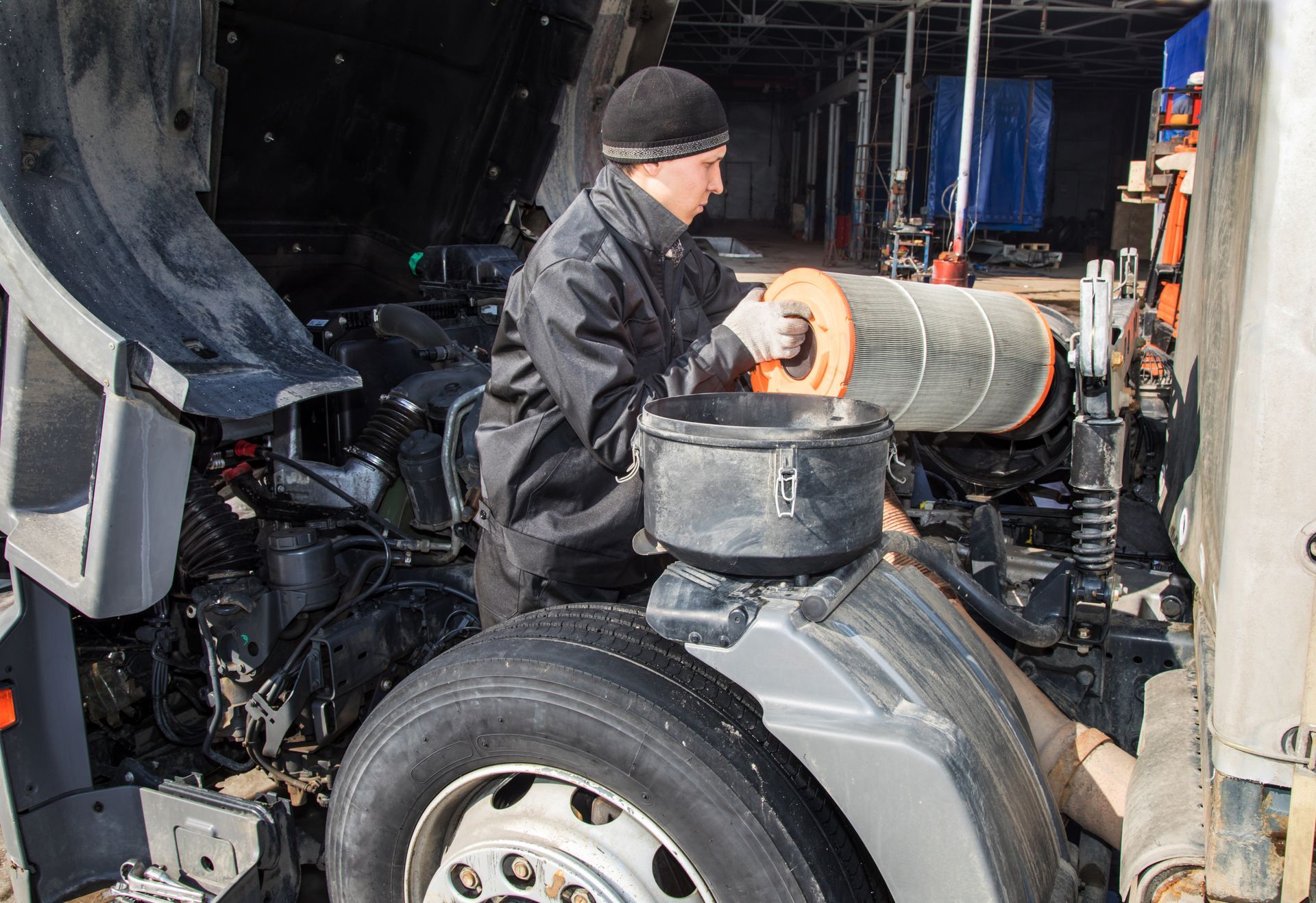 A mechanic in a black jacket and beanie holds a cylindrical truck air filter while working on an open engine bay.