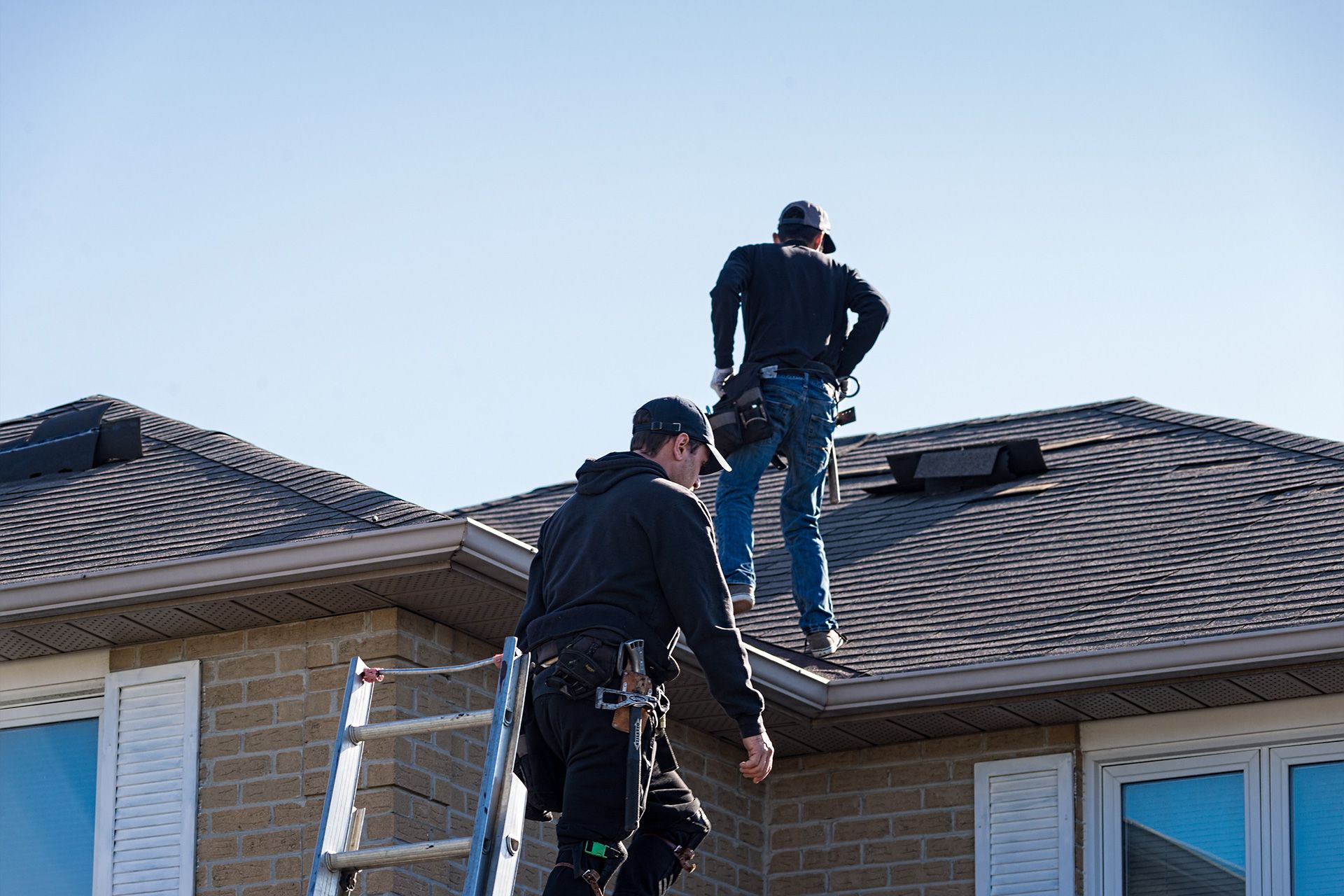 Two workers on a house roof inspecting or repairing shingles under a clear sky