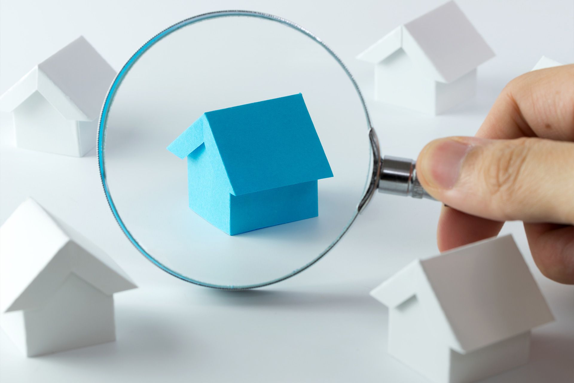 Magnifying glass over a blue house model among white houses, held by a hand