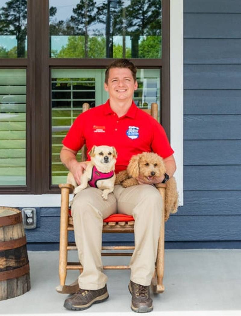 Man in red polo sits on porch with two small dogs in his lap, in front of blue siding and windows- K yle Cobb