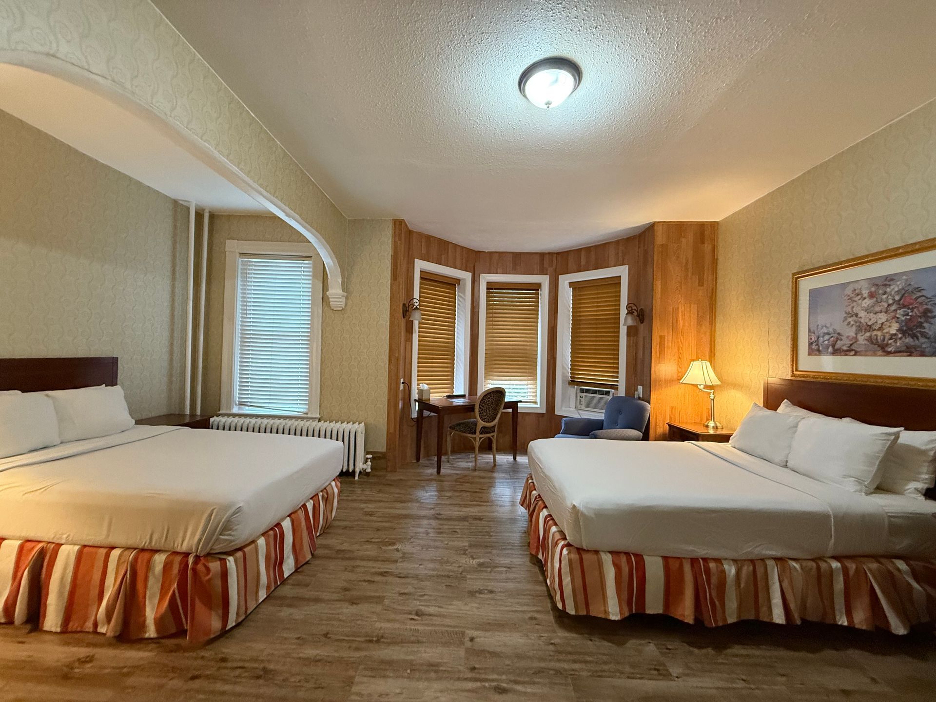 Hotel room with two queen beds, desk, and windows. Beige walls, wood trim, and a light fixture.