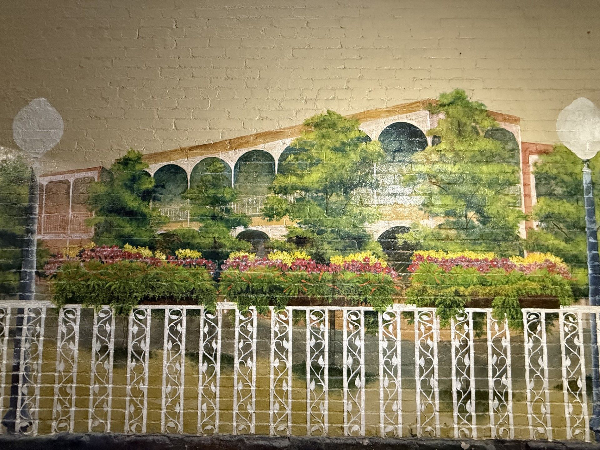 Mural of a building with arched windows, surrounded by green foliage, flowers, and a white decorative fence.
