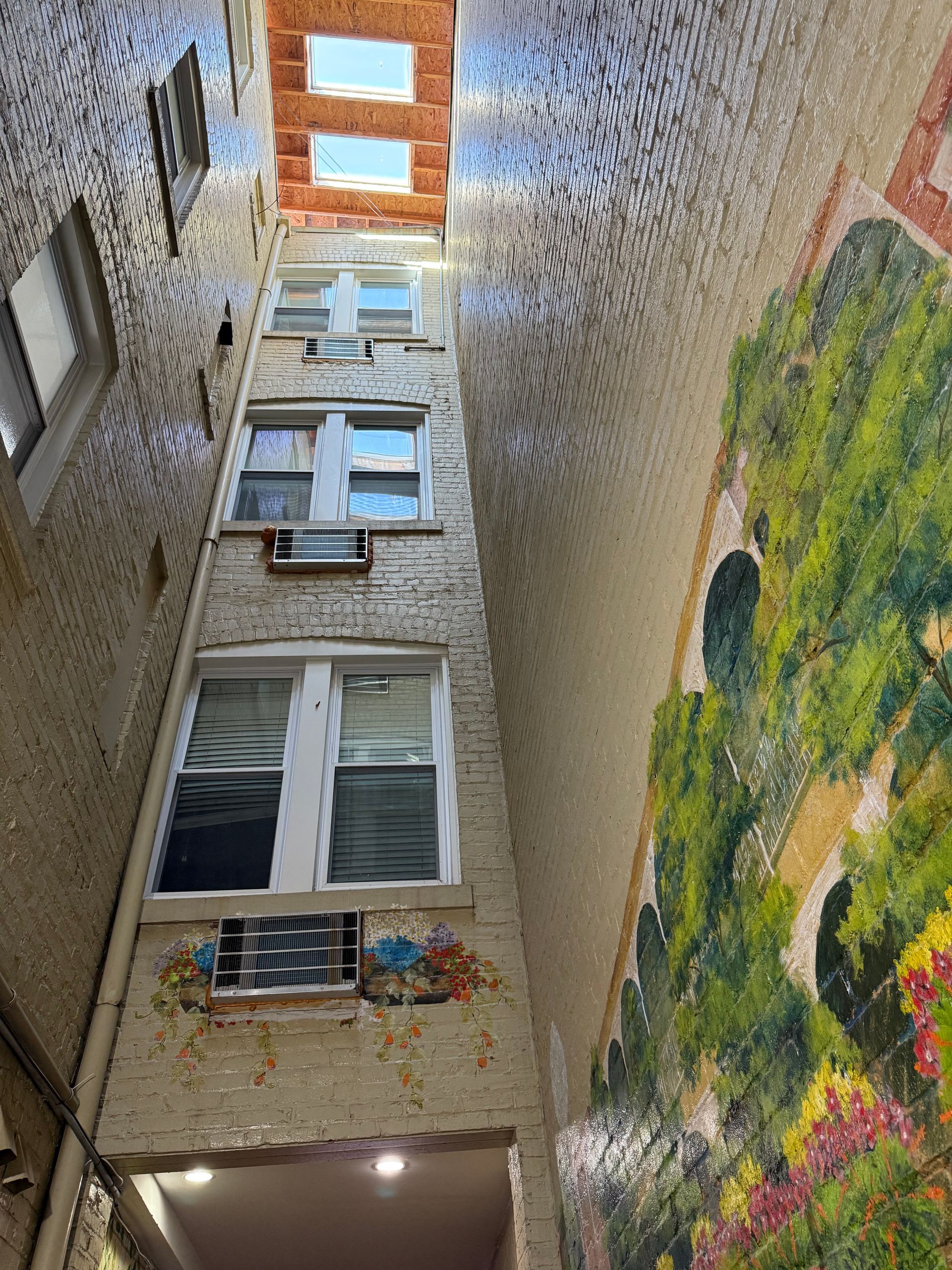 Narrow, multi-story courtyard with windows, air conditioners, and a mural of a garden scene.