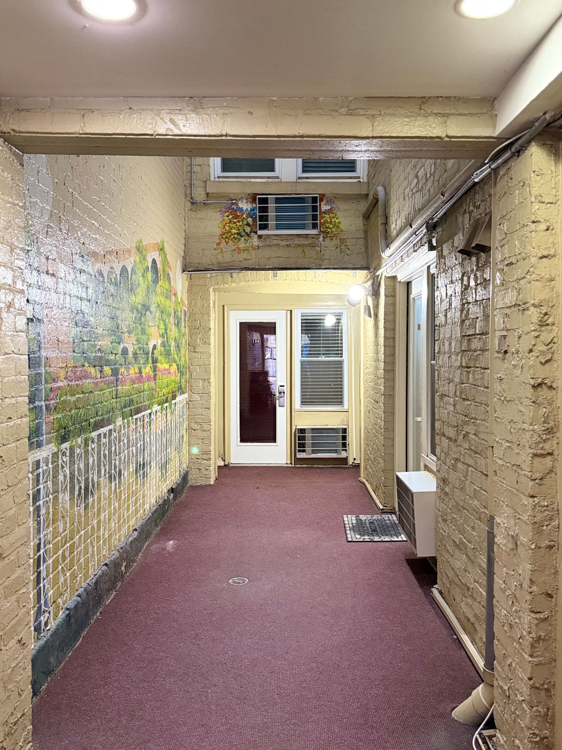 Narrow hallway with mural, doors, and air conditioners; burgundy carpet and tan brick walls.