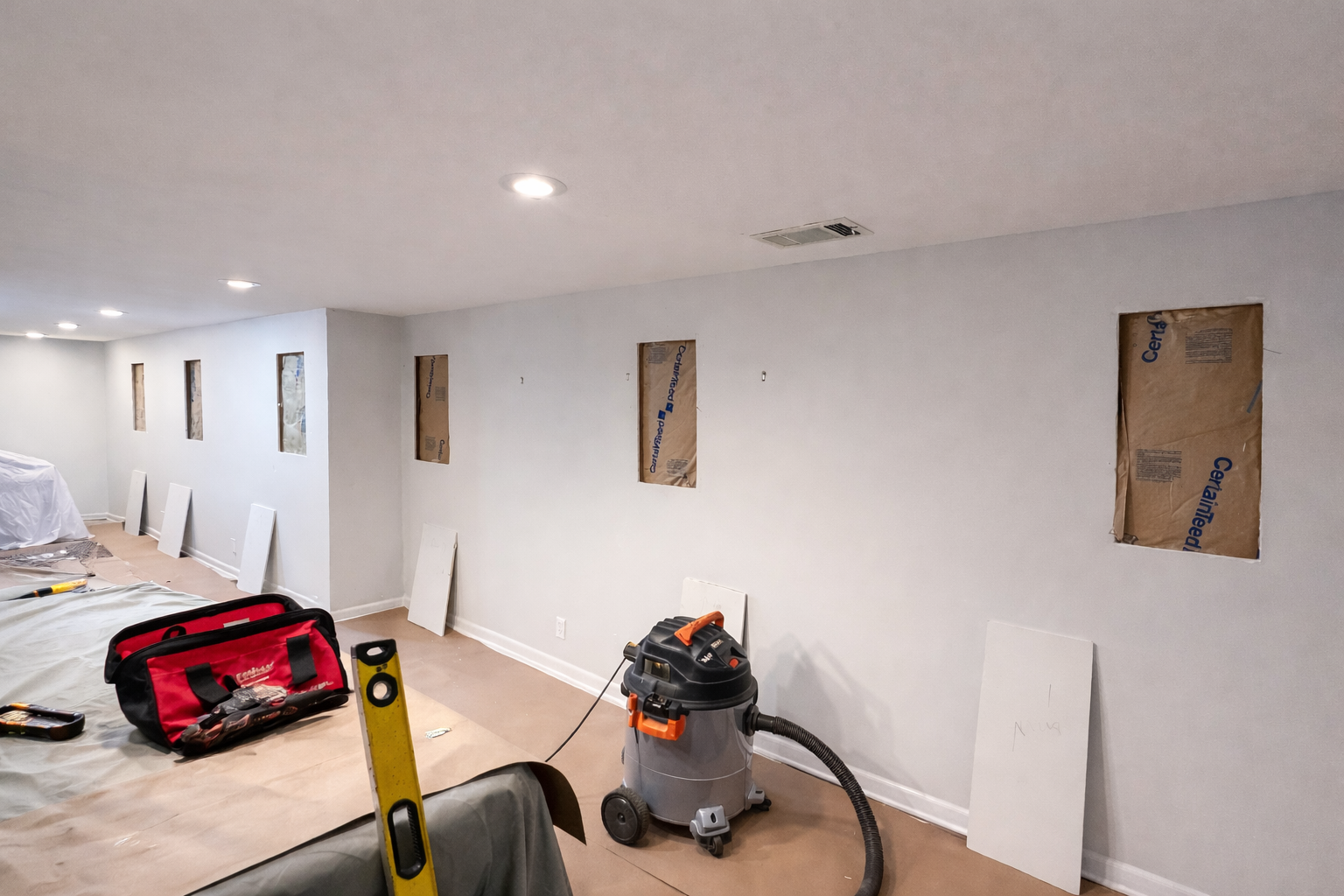 Sump pump system with gray pump, gray PVC pipes in a corner of a room with exposed wooden flooring, and concrete block wall.