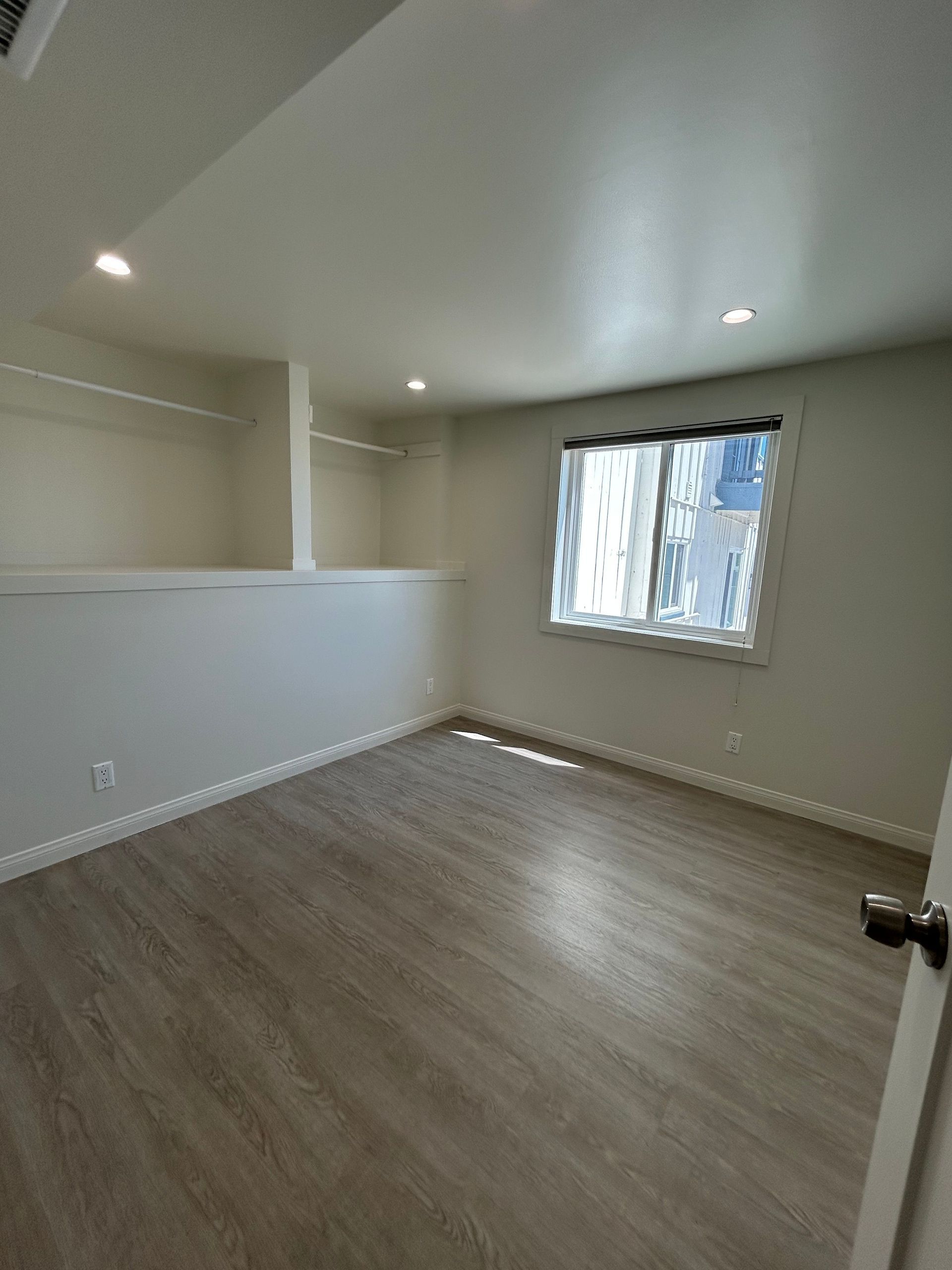Empty beige room with wood-look flooring, recessed lights, and a window with blinds.