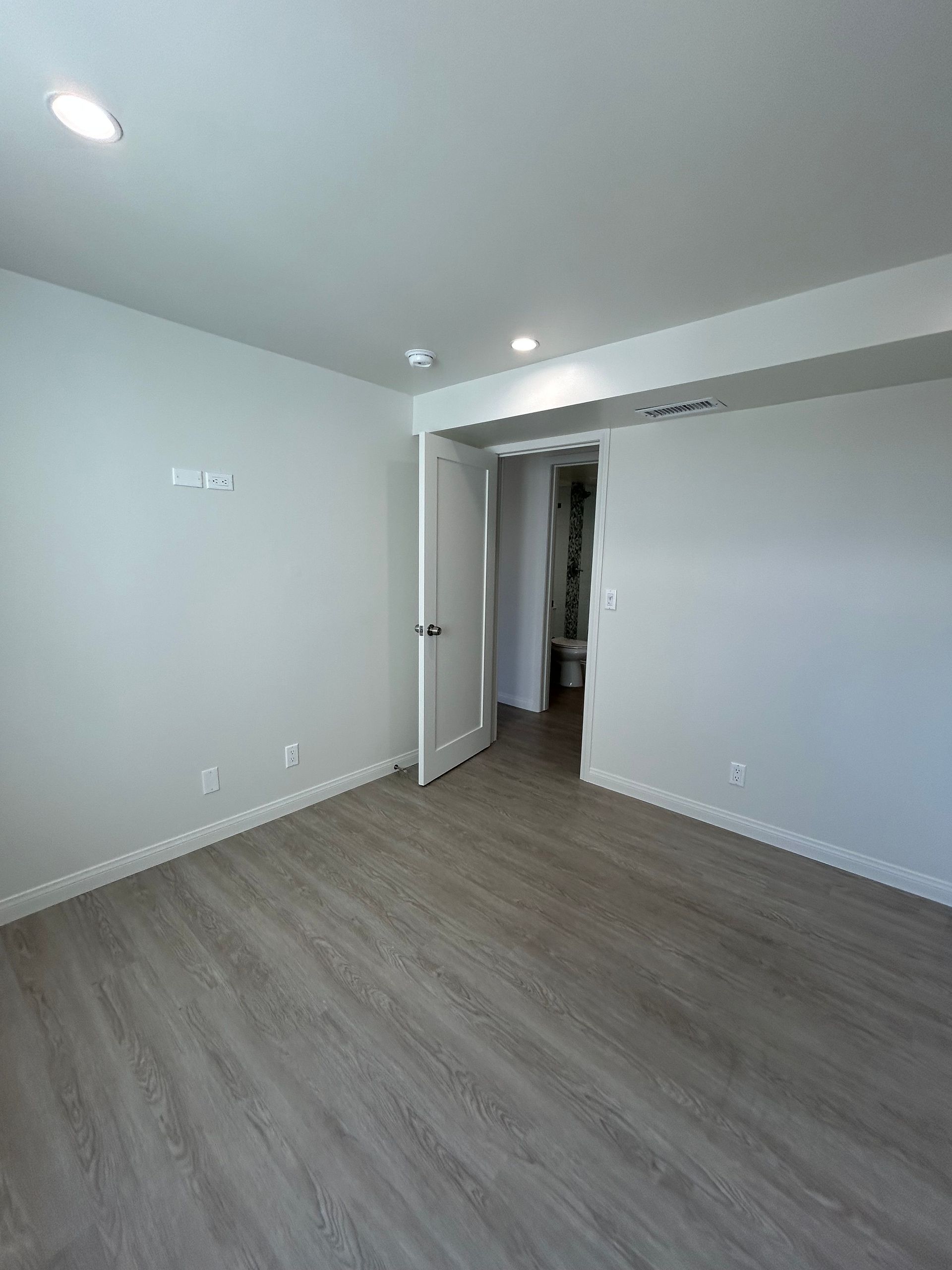 Empty modern room with light gray walls, wood-look flooring, recessed lights, and an open doorway.