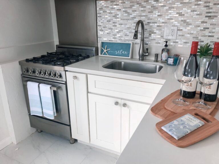 Small modern kitchen with stainless stove, white cabinets, sink, wine bottles, and a wooden tray on the counter