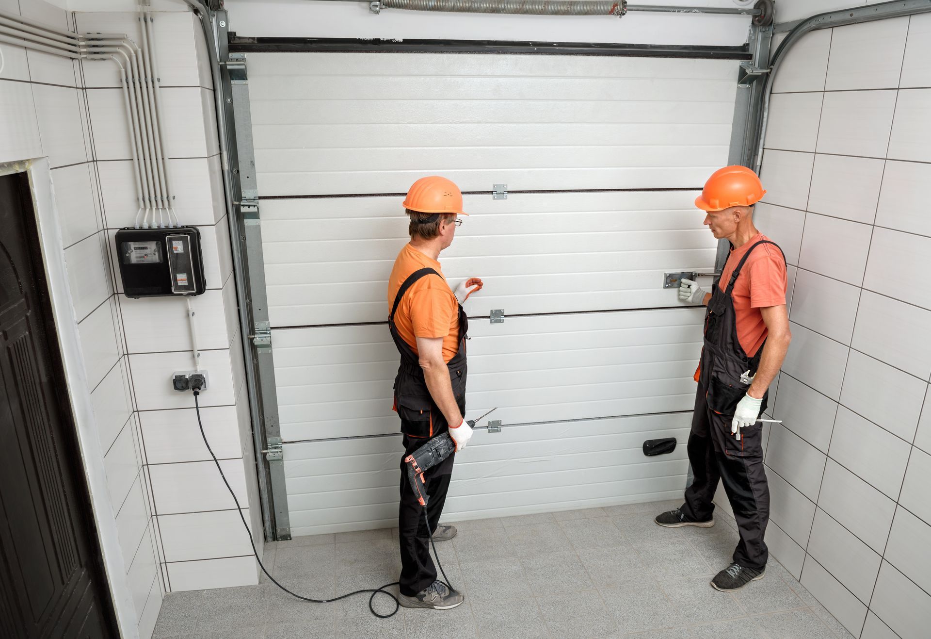 Two workers in orange hard hats and overalls examine a white garage door. Two workers in orange hard hats and overalls examine a white garage door.