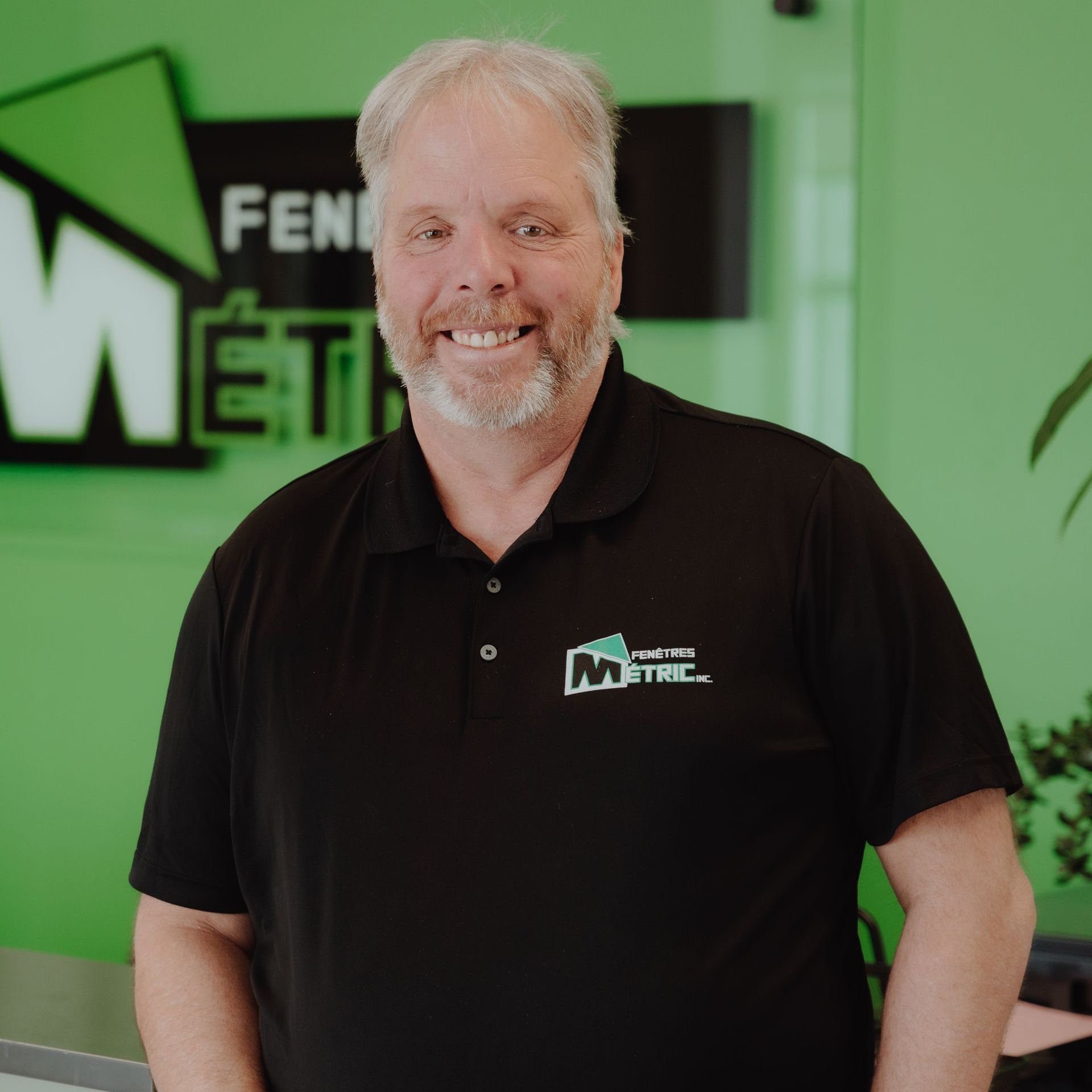 Homme aux cheveux gris et à la barbe en chemise noire avec un logo, souriant devant un mur vert avec une enseigne commerciale