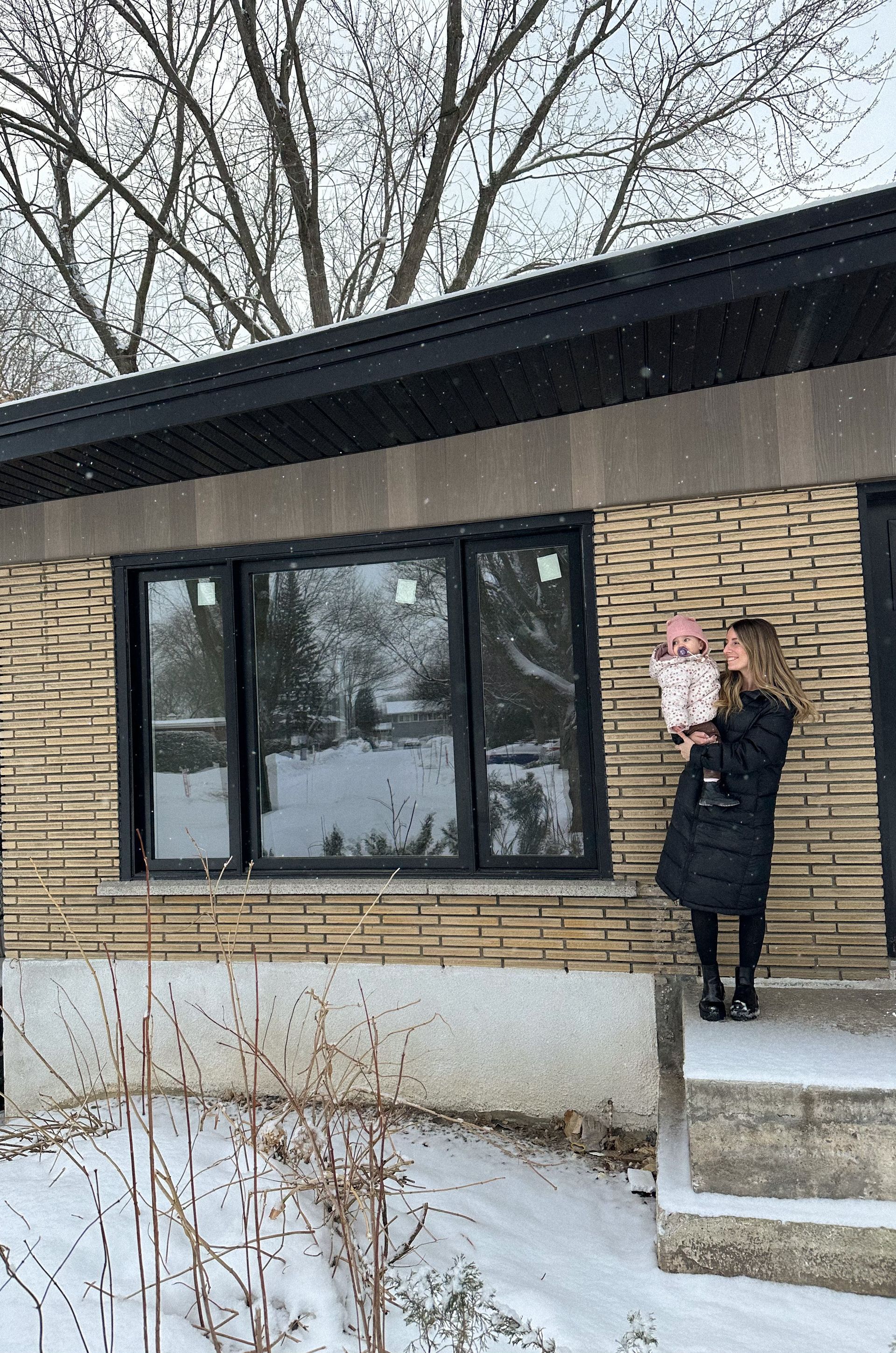 Une femme tenant un bébé à l'extérieur d'une maison en briques avec de la neige au sol.