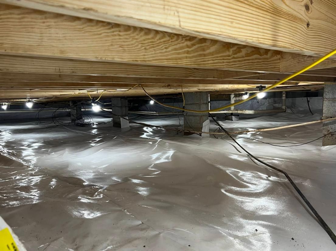 Underneath a house: plastic ground cover, wooden beams, concrete supports, lights, and wiring.