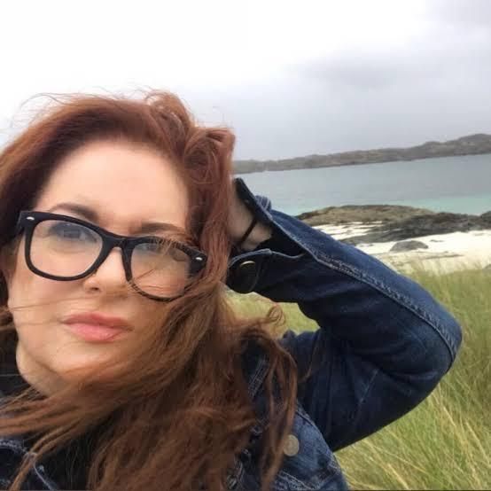 Woman with red hair and glasses poses near a beach on an overcast day.