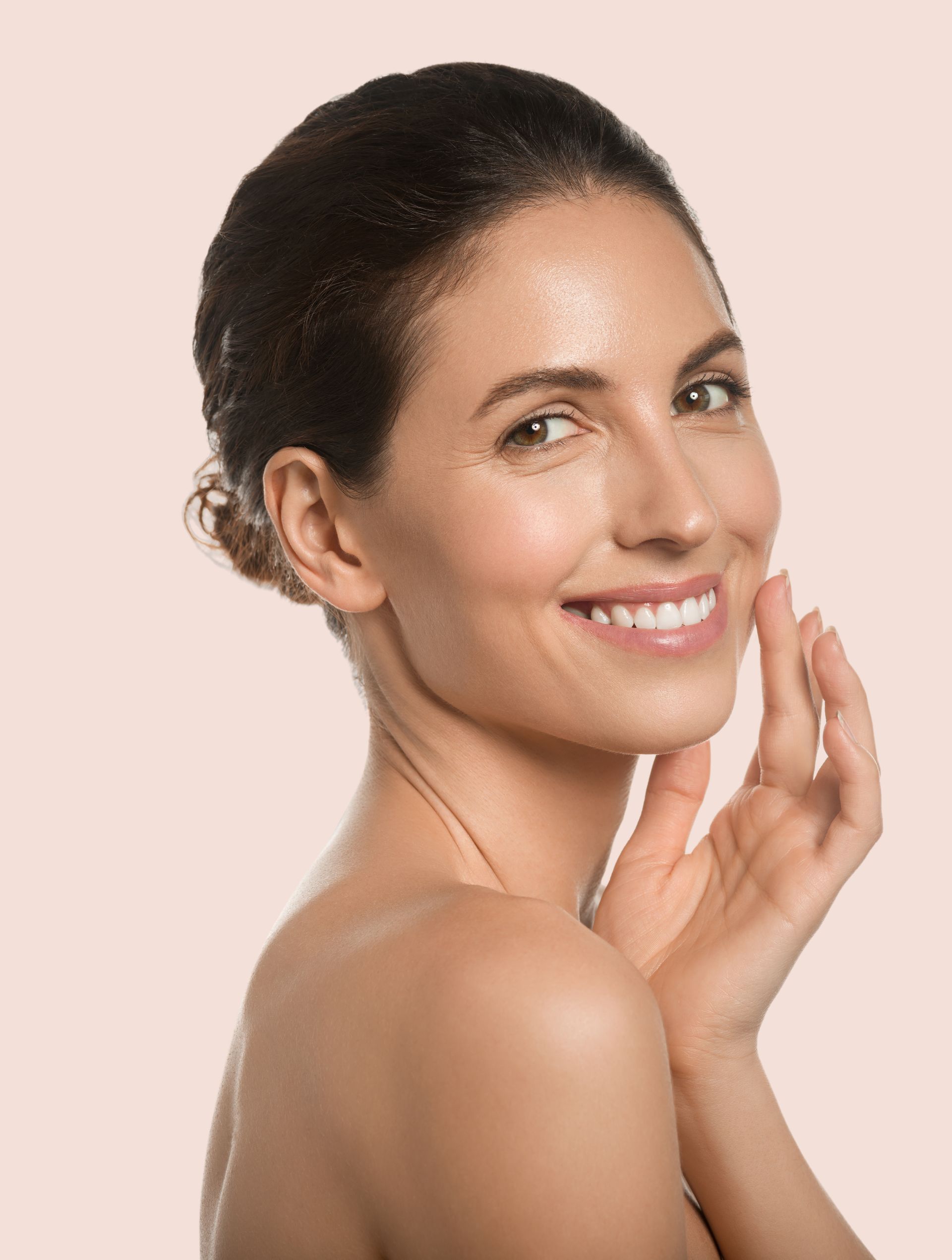 Smiling woman with dark hair on a pink background, touching her face with her hand