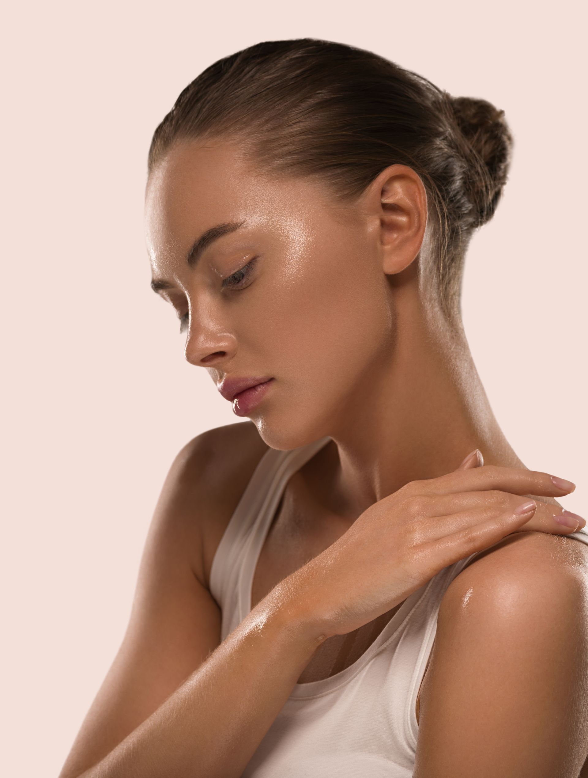 Woman with hair in a bun gently applying lotion to her shoulder against a soft pink background