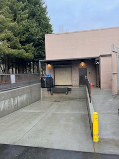 Loading dock with a ramp, building with beige walls, trees in the background, and black bins.
