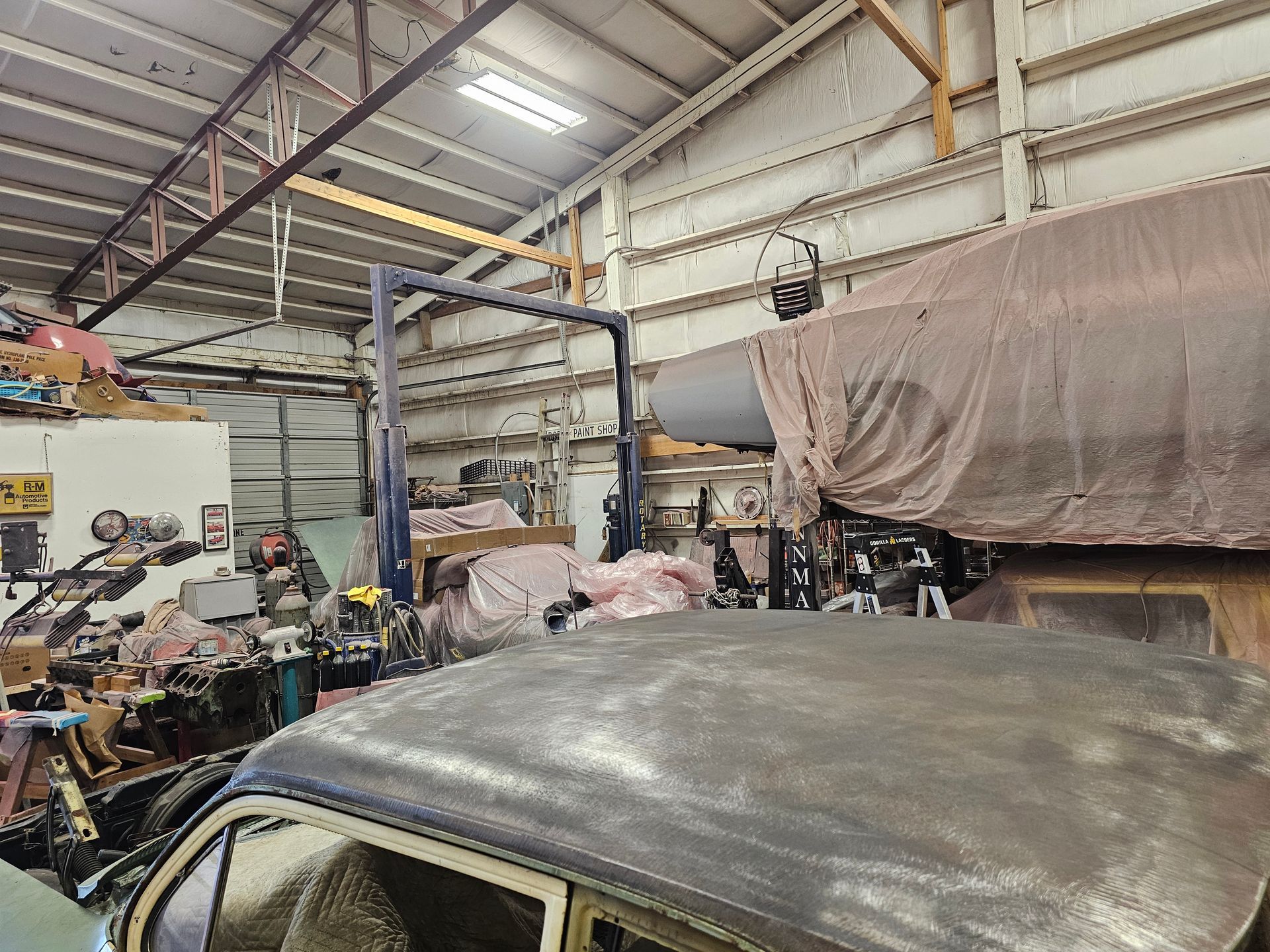 Interior of a cluttered workshop. An old car is in the foreground, other vehicles, tools, and machinery are scattered about.