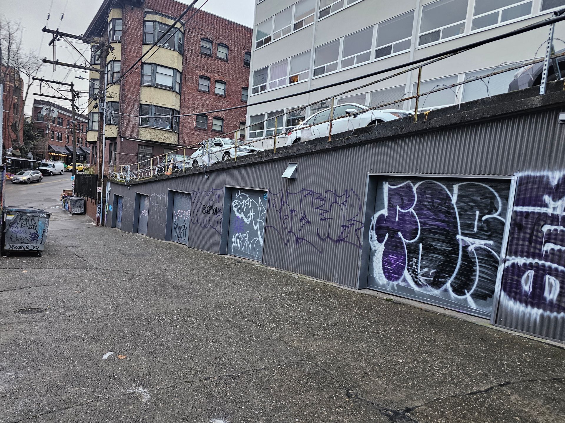Graffiti-covered concrete wall with doorways; street with parked cars and buildings in background.