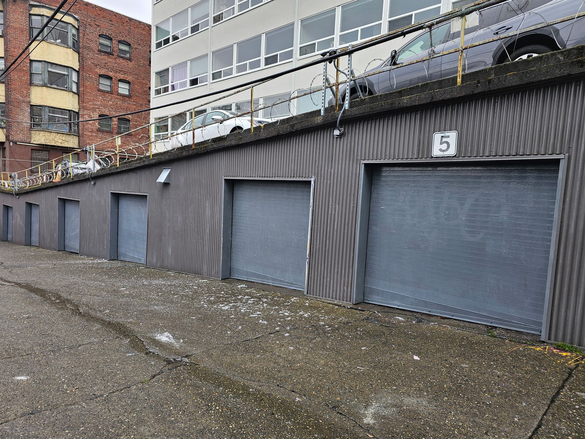 Row of grey storage units along an alley, with a building and cars in the background.