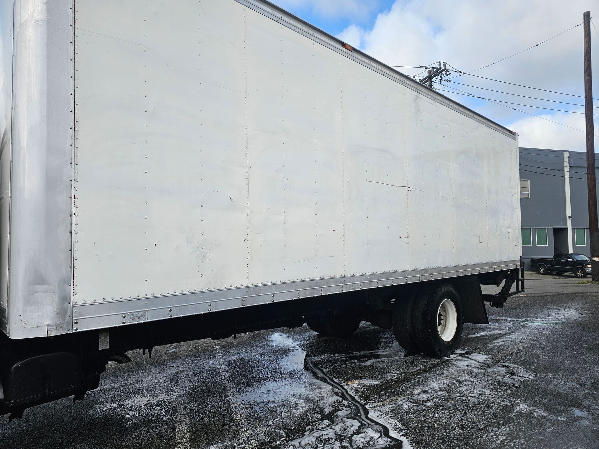 Side view of a white box truck parked on a wet surface with a building and sky in the background.