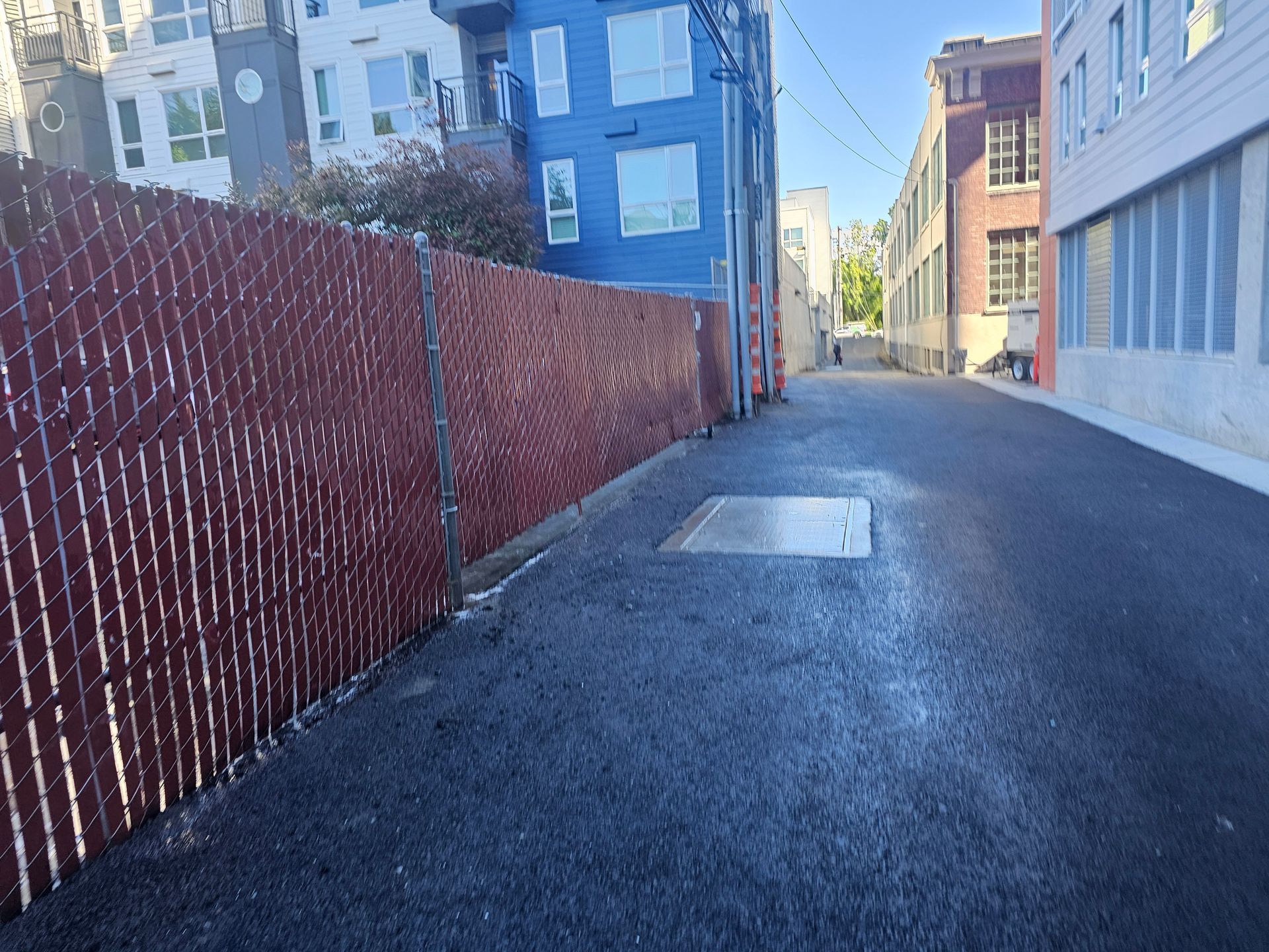 Narrow asphalt alleyway with buildings and a red chain-link fence.